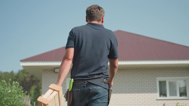 back view of man walking along farm carrying wooden crate and plier while inspecting crops near house roof on sunny day calm rural scene showing agriculture work and crop care