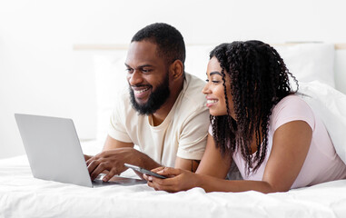 Happy young black husband and wife lie on bed and watch video on computer in free time in bedroom...