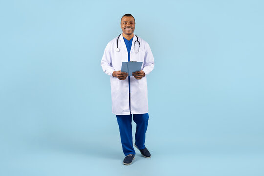 African American male doctor standing with clipboard in lab coat, smiling at camera in studio. Portrait of confident young professional physician against blue background