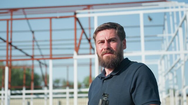 Bearded man in dark polo near metal frame structure folds pneumatic rifle, break barrel action with careful safety check, focused squint under bright summer daylight, rural yard setting