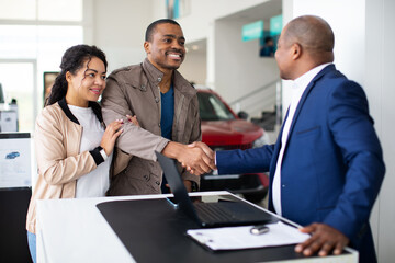 Smiling African American couple shaking hands with car dealer at desk, celebrating successful purchase agreement, lifestyle moment of auto shopping, finance and trust