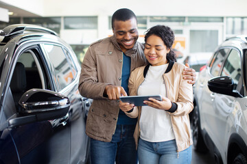 Happy African American couple using tablet in car showroom, smiling while reviewing auto purchase details, lifestyle concept of technology, business, transport and finance