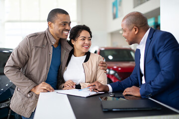 Happy African American couple smiling with car dealer at desk in showroom, reviewing contract papers, lifestyle and transport concept of business, purchase and ownership
