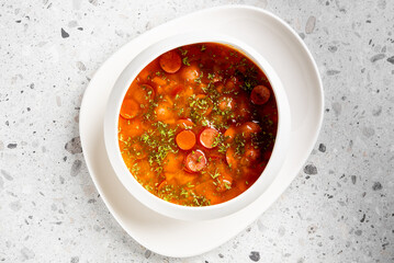 Hearty tomato-based soup with beans, carrots, sausage slices, and herbs served in a white bowl on a...