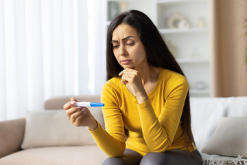 Thoughtful pregnant woman in yellow top holding pregnancy test, sitting on sofa at home. Emotional prenatal lifestyle moment with anticipation and concern