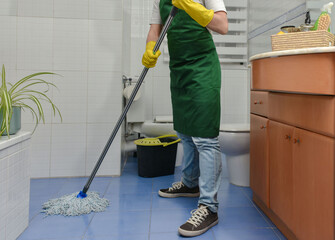 Cropped view of a person in a green apron and yellow cleaning gloves mopping the bathroom floor...