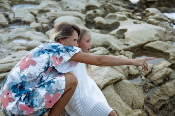 Mother and daughter enjoying time by the sea. Smiling woman and young little girl sitting on rocks at the seaside, bonding and pointing into the distance.