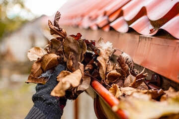 A gloved hand clears the gutter of a red tile roof of fall leaves, preventing clogs and water damage