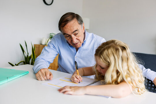 Young little cute girl doing homework with her grandfather at home. Old man helping her daughter with school tasks. Education and family concept.