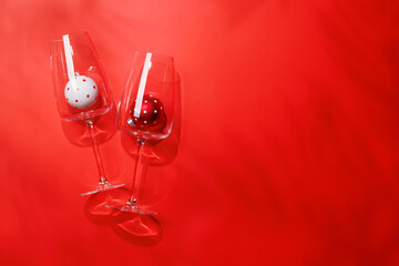 Wine glasses with Christmas baubles inside on red background, mockup for holiday card or invitation