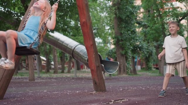 playful girl laughs out loud on wooden swing with chain handle as her playmate pushes from behind in lush green park, capturing candid joy motion blur and sibling fun in warm evening light