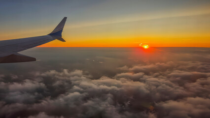 An airplane wing flies above the clouds at dawn with the rising sun	
