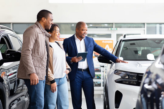 Smiling African American couple with car dealer examining new SUV in modern showroom, lifestyle and transport moment while considering purchase options for vehicle.