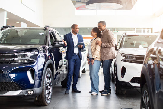 Smiling African American couple with car dealer in showroom, looking at new SUV, discussing purchase details, lifestyle and business moment of modern auto shopping