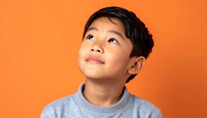 A young boy with dark hair smiles sweetly, looking upwards against an orange background. He wears a grey shirt and appears thoughtful, hopeful, or perhaps curious