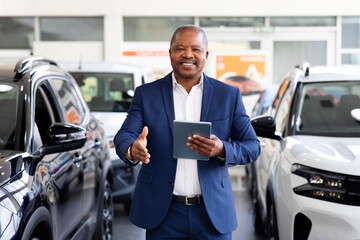 Smiling African American car dealer in blue suit greeting customers with handshake inside showroom, confident professional lifestyle moment in modern auto sales business
