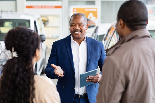 African American car dealer smiling while speaking with young couple in showroom, friendly consultation about purchase and finance, lifestyle moment of auto sales