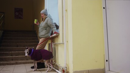 Woman in grey beanie and green puffer jacket holding cup stands beside whippet in purple coat with toy around neck, both waiting in tiled indoor hallway near stairs in winter, casual urban moment