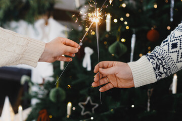 Happy New Year! Burning sparklers in hands on background of stylish decorated christmas tree and fireplace with golden lights. Glowing fireworks in hands, holiday celebration