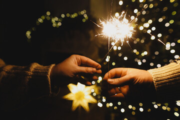 Hands holding fireworks against christmas tree lights bokeh in evening cozy living room. Happy New...