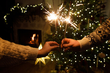 Hands holding fireworks against christmas tree lights in evening cozy living room. Happy New Year eve! Atmospheric family celebration with burning sparklers in hands close up