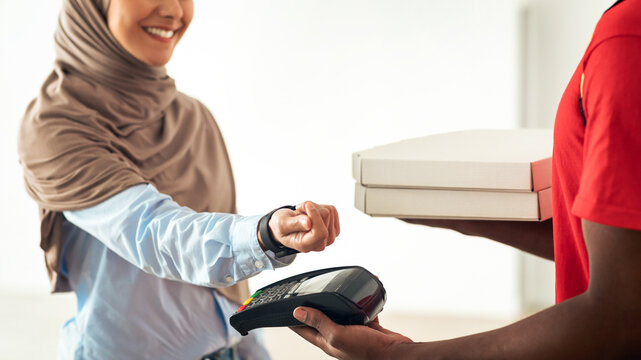 Fast Food Delivery, Easy Payment. Closeup of black male courier wearing red uniform holding Point of Sale terminal and pizza boxes, smiling muslim woman in hijab paying using smart watch