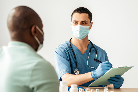Collects data of patient and preparation for immunization in clinic. Young doctor in protective mask and african american male patient notes questionnaire in medical office with vials and drugs
