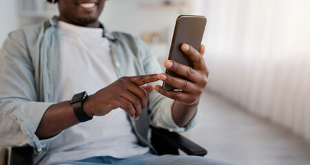 Audio app, gadgets at home, relax at home and enjoy music. Happy adult african american male disabled in wheelchair in wireless headphones with smartphone listen song in living room interior, cropped