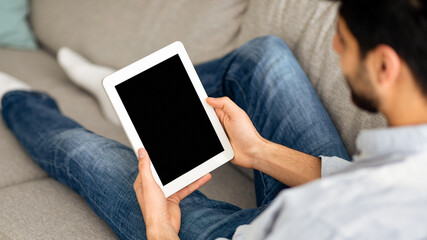 Advertisement concept. Millennial arab man using digital tablet with blank black screen, browsing website or showing new app, sitting on sofa at home. Mockup for your design