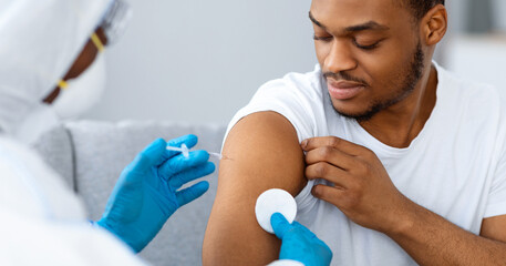 Vaccination at home concept. Nurse in protective suit making COVID-19 vaccine for black guy at home, closeup. African american young man receiving immunization shot during coronavirus pandemic