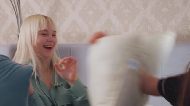 Blonde girl laughs with hand near face while sitting on bed during lighthearted pillow fight, her expression shows excitement and happiness in cozy bedroom