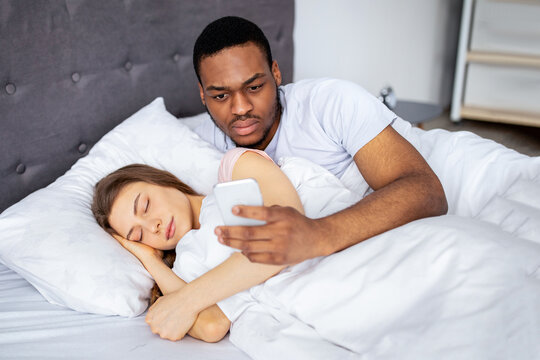 Jealous black husband checking his wife's smartphone, reading correspondence while she is asleep in bed at home. African American guy suspecting partner's infidelity. Marriage, relationship problems