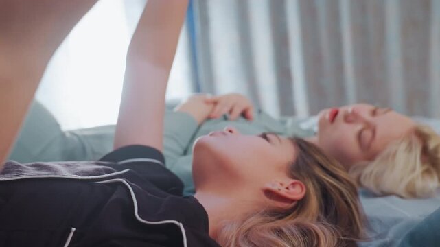 Close up of two girls lying on bed in relaxed indoor atmosphere, one with arm raised holding object possibly phone, other lady resting, with natural light coming through curtain in background