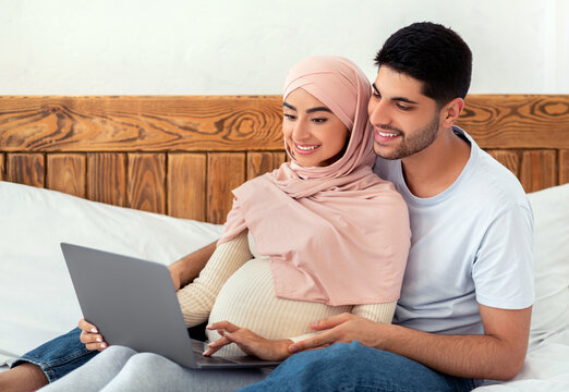 Cheerful pregnant arab couple using laptop computer, watching movie together while resting on bed, expecting muslim lady spending time with her husband, empty space - Powered by Adobe