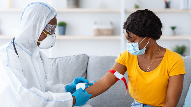 Nurse in protective suit taking blood sample from vein for female patient at home, african american doctor checking young lady blood for coronavirus antigens or making general checkup