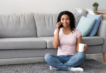 Fototapeta premium Free time, rest and calls to friend on phone, coffee break at home. Satisfied millennial pretty african american woman talking on smartphone, hold cup of hot drink in minimal living room interior
