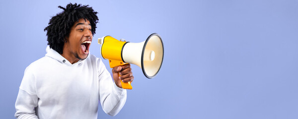 Cool news. Agitated African American teen guy screaming into megaphone over blue studio background....