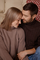 Young Caucasian couple, man and woman in warm sweaters, sitting close, hugging gently, front view. Indoors, cozy room with soft sofa. Concepts: love, intimacy, warmth, couple bonding. 