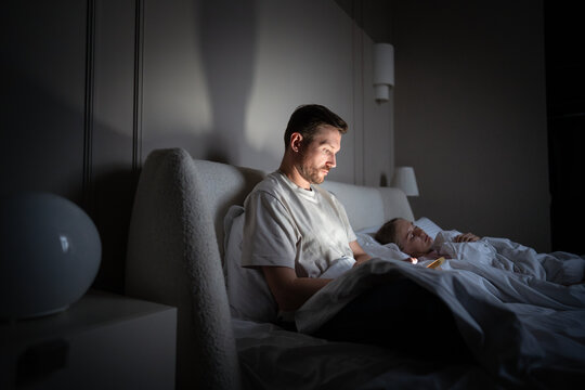 Unfaithful husband scrolls smartphone, engages in social media chats in bed at night. Wife sleeps unaware as husband strays fixated on phone, digital addiction and demanding job, affair and cheating