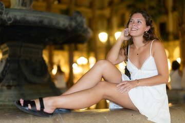 Woman in a white dress sits near a fountain in a night square in Barcelona, she enjoys a summer...