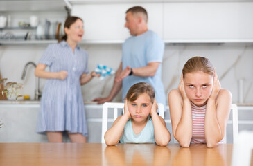 Frightened and frowning children sit at kitchen table during mom and dad quarrel. Behind children,...