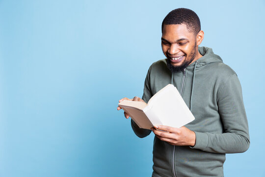 African american bookworm engrossed in his reading leisure time against blue background, narrating a novel story aloud for the audience. Young casual adult relaxing with literature.
