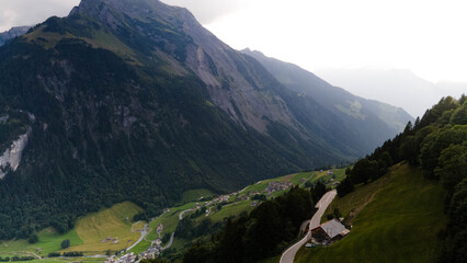 Swiss Alps with houses, winding road, clouds, and overcast sky, captured by drone. Schweizer Alpen mit Häusern, kurviger Straße, Wolken und bedecktem Himmel, aufgenommen mit Drohne.