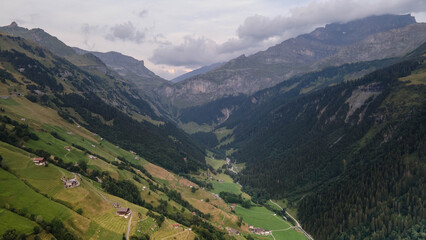Obraz premium Swiss Alps with houses, winding road, clouds, and overcast sky, captured by drone. Schweizer Alpen mit Häusern, kurviger Straße, Wolken und bedecktem Himmel, aufgenommen mit Drohne.