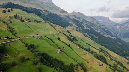 Obraz premium Swiss Alps with houses, winding road, clouds, and overcast sky, captured by drone. Schweizer Alpen mit Häusern, kurviger Straße, Wolken und bedecktem Himmel, aufgenommen mit Drohne.