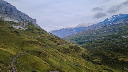 Swiss Alps with houses, winding road, clouds, and overcast sky, captured by drone. Schweizer Alpen mit Häusern, kurviger Straße, Wolken und bedecktem Himmel, aufgenommen mit Drohne.