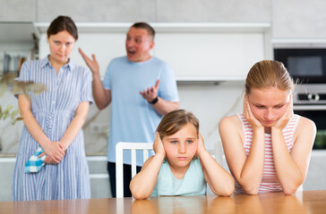 Frightened and frowning children sit at kitchen table during mom and dad quarrel. Behind children,...