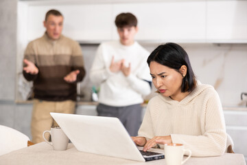 Young guy and adult man quarreling with young woman working on laptop in kitchen at home..