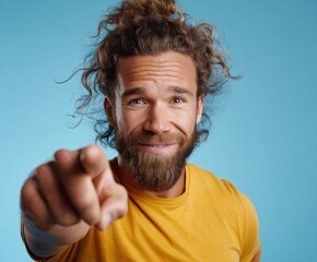 A man with curly hair and a beard, wearing a mustard-yellow t-shirt, points directly at the viewer with a friendly, engaging expression against a vivid light-blue background.
