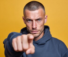 Close-up portrait of a man in a dark blue hoodie, pointing directly at the viewer, against a bright yellow background.